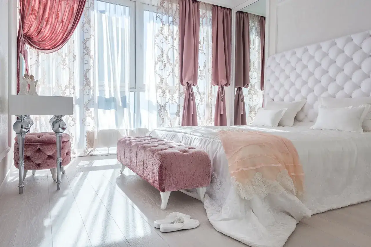 Elegancka sypialnia w stylu angielskim z pikowanym białym zagł&oacute;wkiem, r&oacute;żowymi pufami i zasłonami.