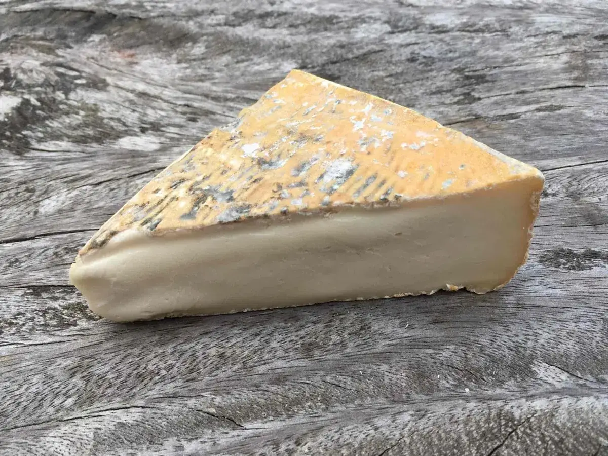 Fetta di formaggio taleggio con crosta rugosa e pasta chiara e compatta, adagiata su un tavolo di legno grezzo.