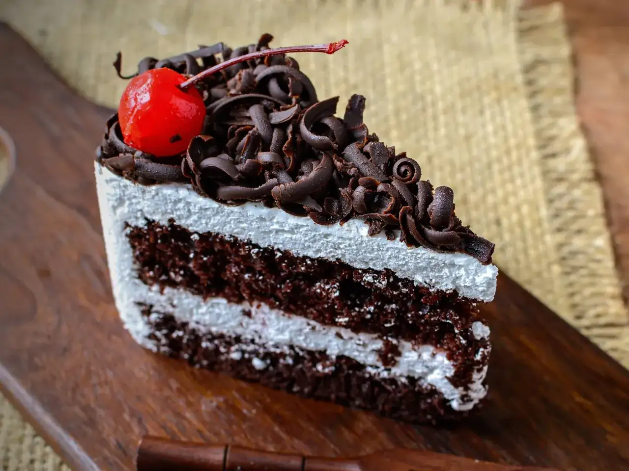 Una fetta di foresta nera torta, con panna montata, riccioli di cioccolato e una ciliegina rossa, su un tagliere di legno.