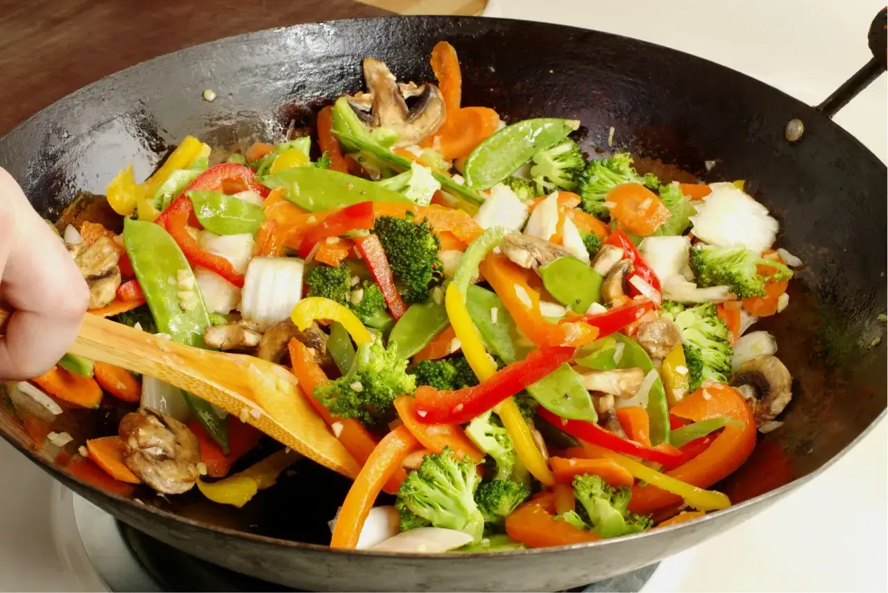 Un colorido salteado de verduras con brócoli, pimientos, zanahorias y guisantes, removido con una espátula de madera en un wok.