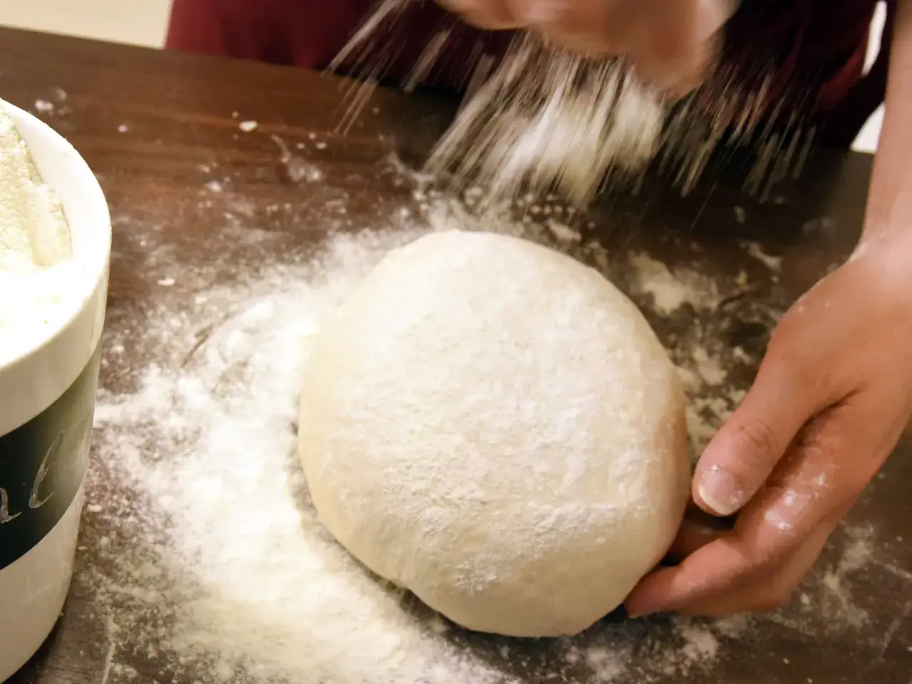 Eine Hand streut Mehl &uuml;ber einen Teigballen. Nebenan steht eine Sch&uuml;ssel mit self-raising flour deutsch.