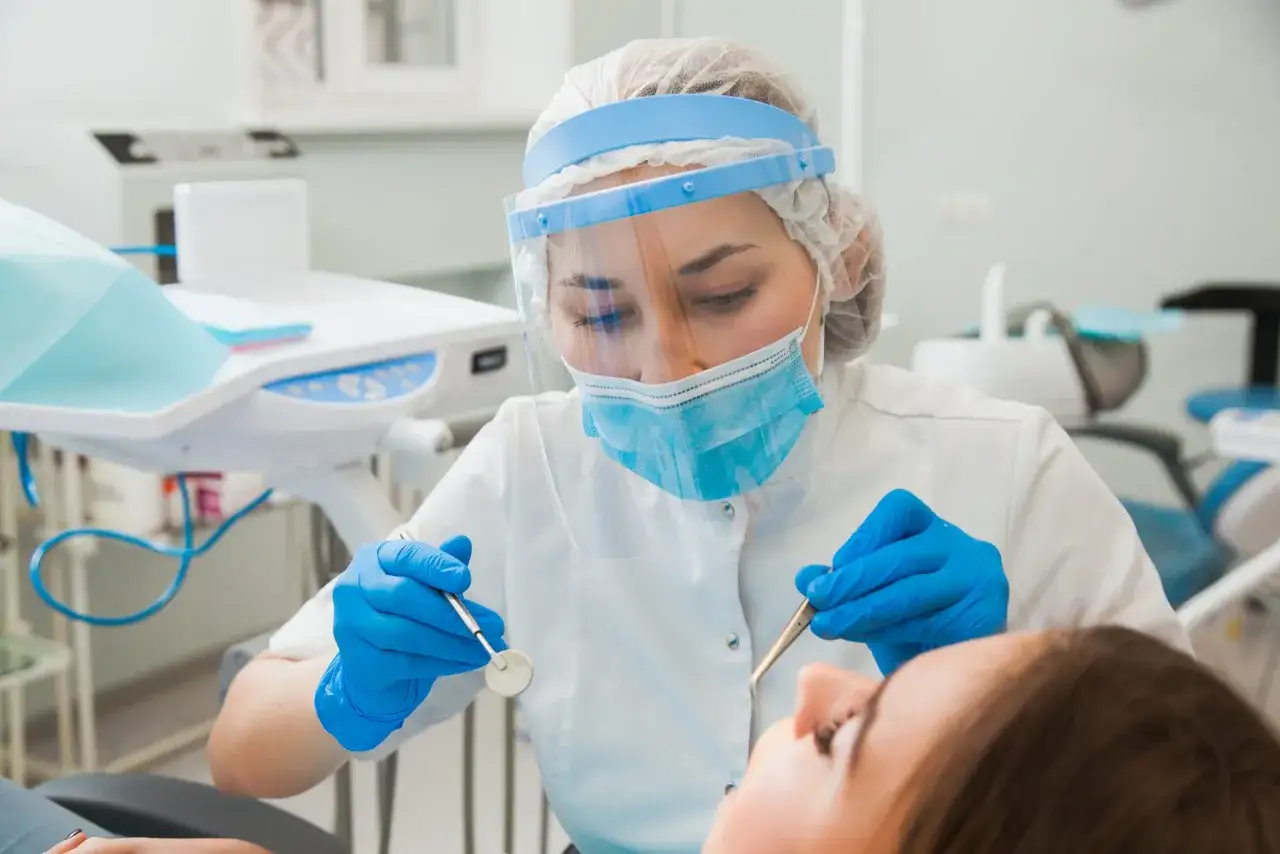 Dentystka w masce i przyłbicy bada zęby pacjentki. Nawet przy przeziębieniu, wizyta u dentysty jest ważna.