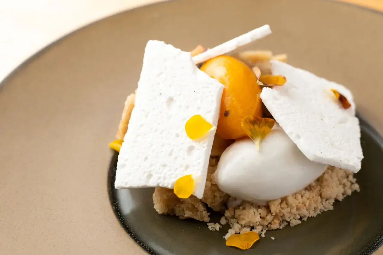 Ein kunstvolles veganes Kokos-Dessert mit Kokos-Eis, Baiser-Splittern, einer orangefarbenen Frucht und Blütenblättern.