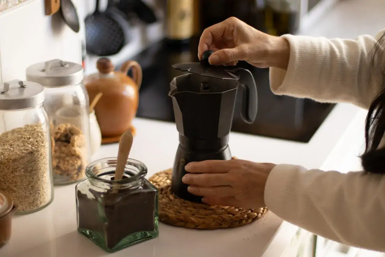 Manos preparando un caf&eacute; con alcohol en cafetera italiana. Ingredientes como avena y caf&eacute; molido a la vista.