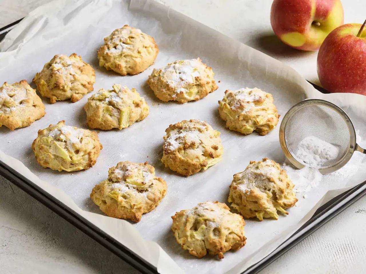 Saftige Apfelkekse, frisch gebacken und mit Puderzucker bestäubt, liegen auf einem Backblech. Daneben zwei Äpfel und ein Sieb mit Puderzucker.