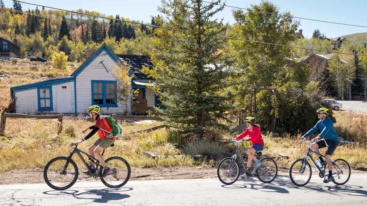 Three cyclists enjoy a scenic ride on a paved road past an old house. The lead cyclist on a mountain bike shows that you can indeed ride a mountain bike on the road.