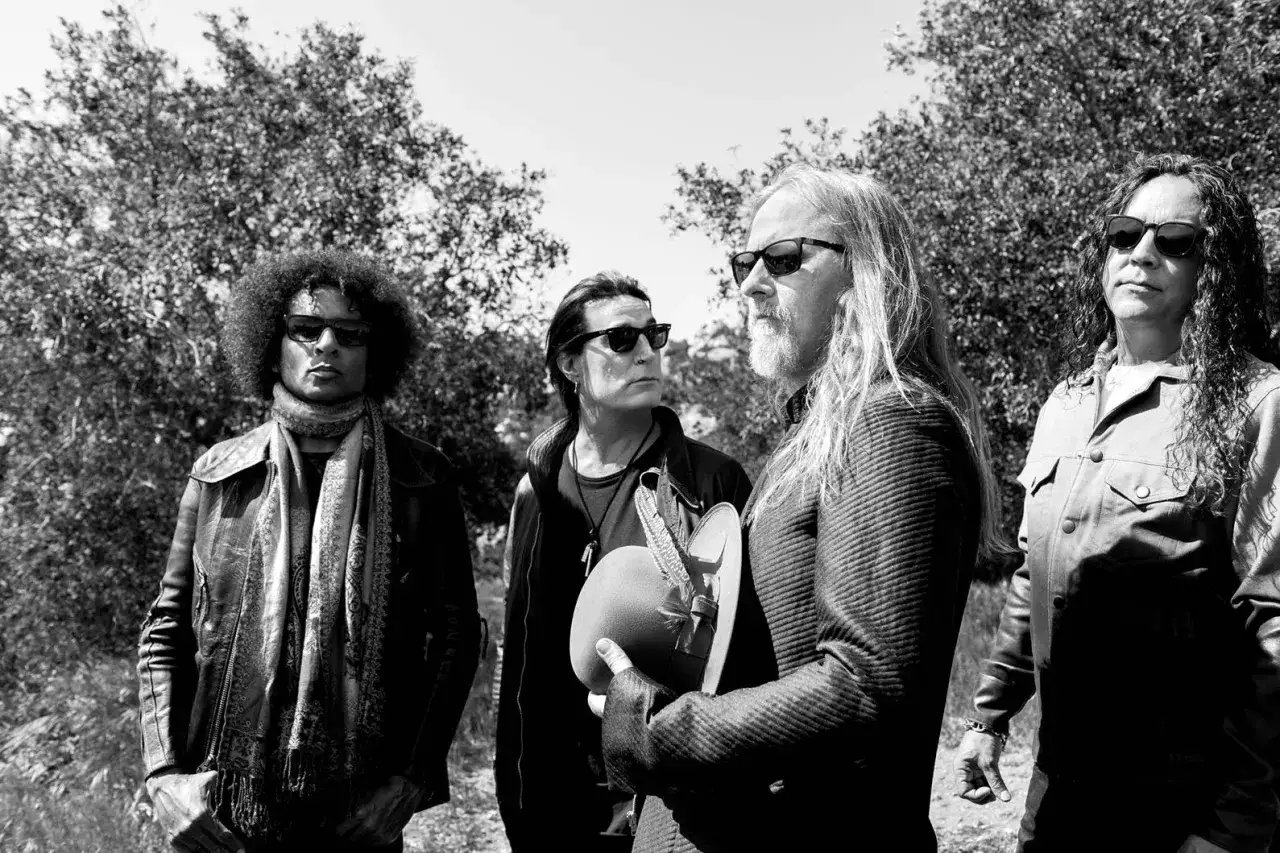 Członkowie Alice in Chains w okularach przeciwsłonecznych, stojący na tle drzew. Jeden z nich trzyma ozdobne nakrycie głowy.