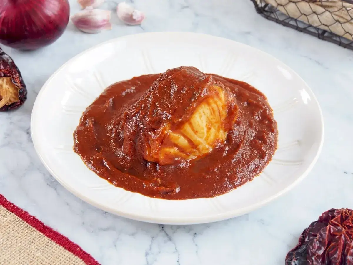 Un delicioso bacalao a la vizca&iacute;na en un plato blanco, con pimientos secos y cebolla morada de fondo.