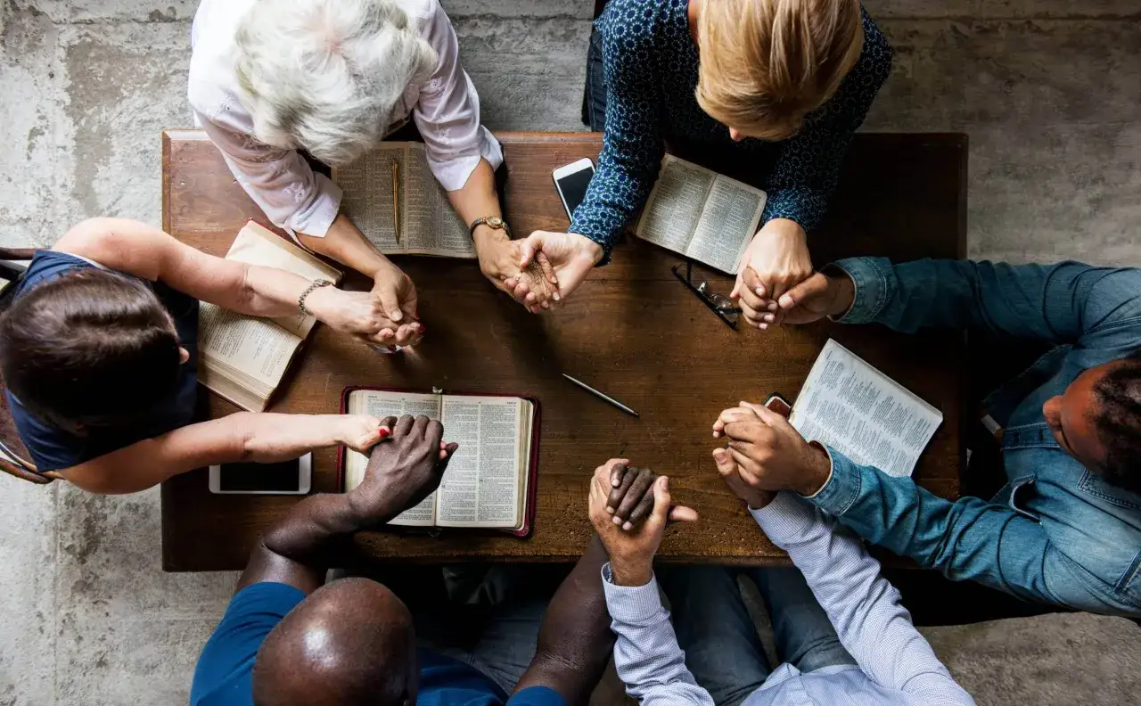 A diverse group of people hold hands around an open Bible, their heads bowed in prayer. This shared moment reflects their statement of faith and unity.