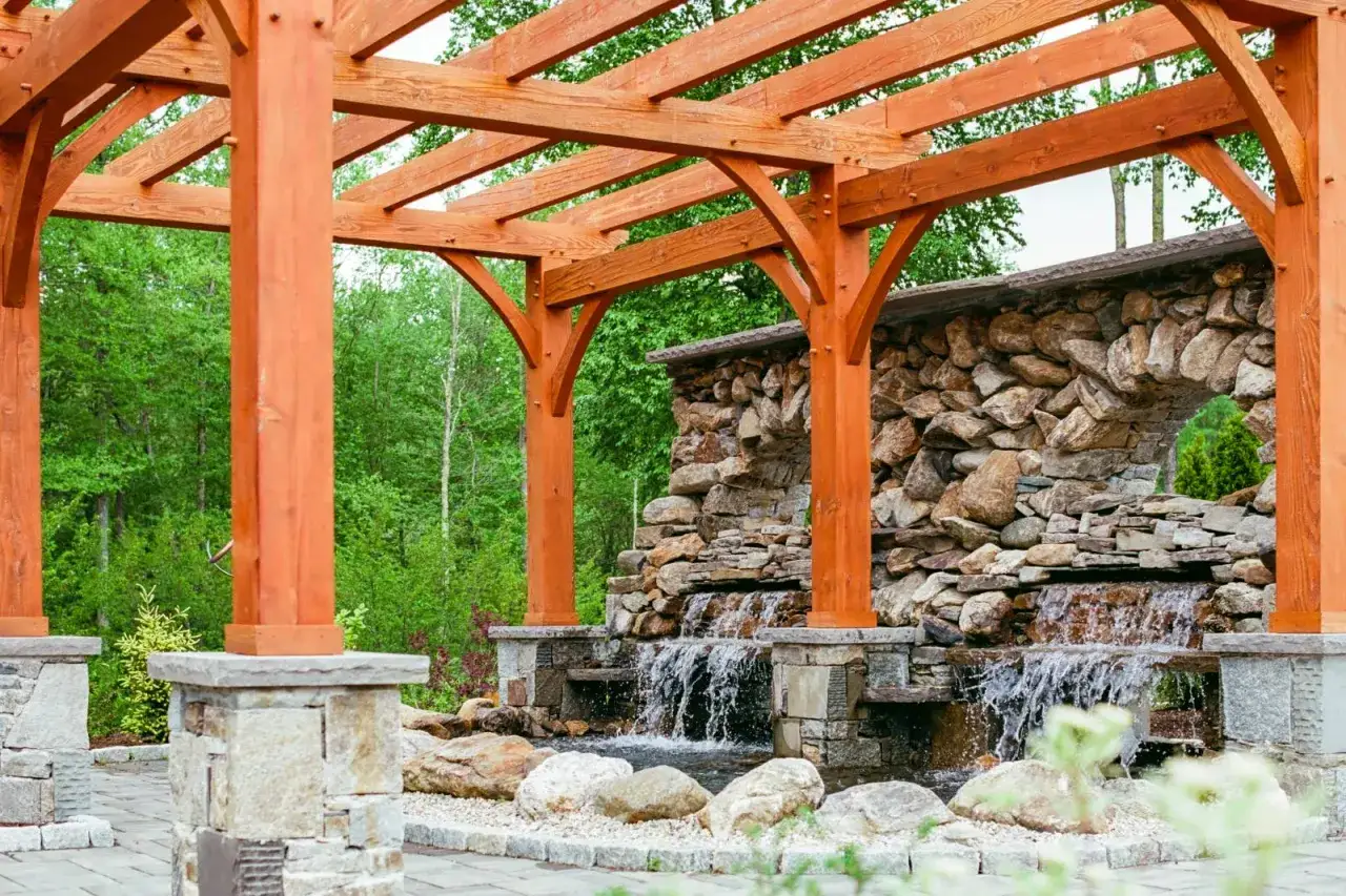 A beautiful wooden pergola frames a cascading stone waterfall, offering inspiration for how to build a pergola in a natural setting.