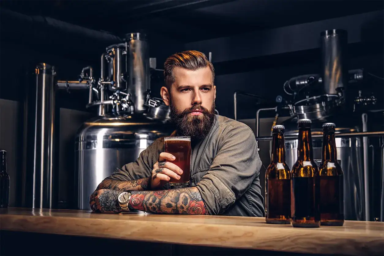 Brodaty mężczyzna z tatuażem na ramieniu trzyma kufel piwa. Obok stoją butelki z alkoholem.