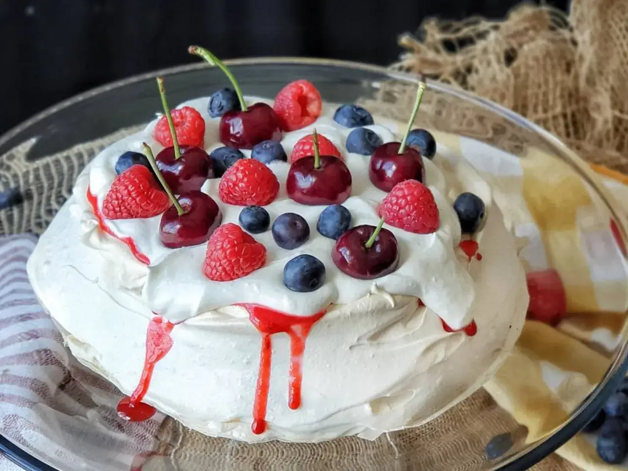Deliciosa pavlova receta original, coronada con cerezas, frambuesas y arándanos frescos, con un toque de sirope rojo.