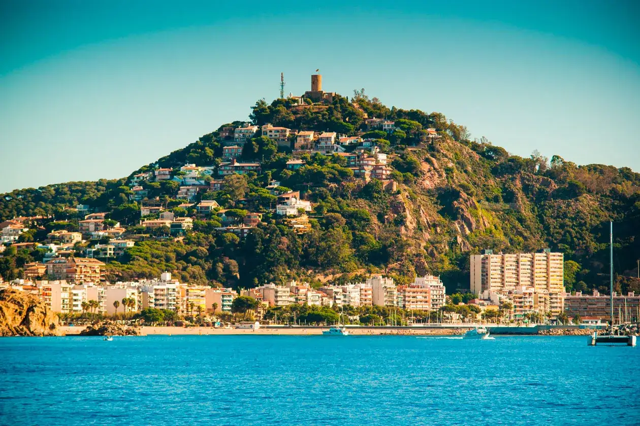 Vista de la costa con casas en la ladera y el **castillo de Blanes** en la cima. El mar azul y los barcos completan la escena.