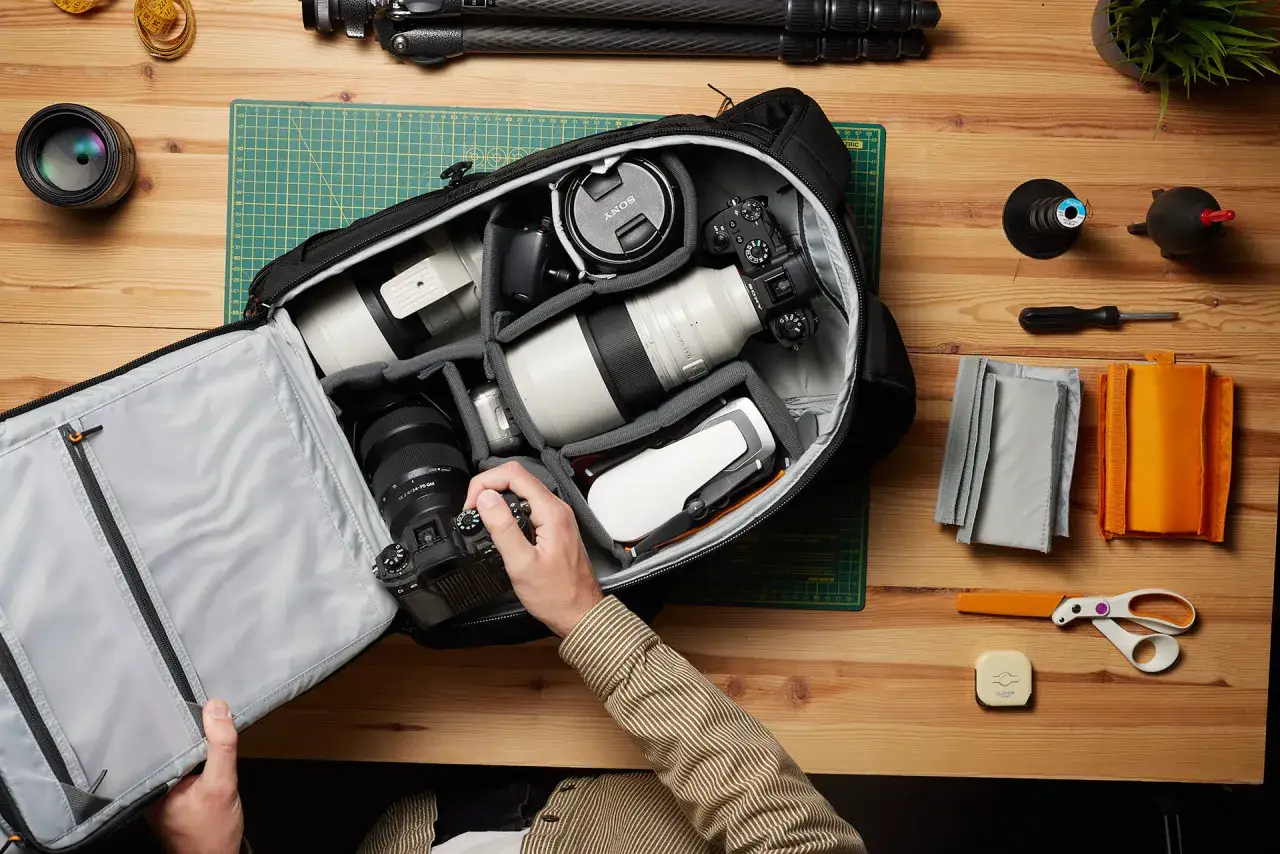 A photographer demonstrates how to pack a camera bag, carefully placing lenses, a drone, and camera body into padded compartments.