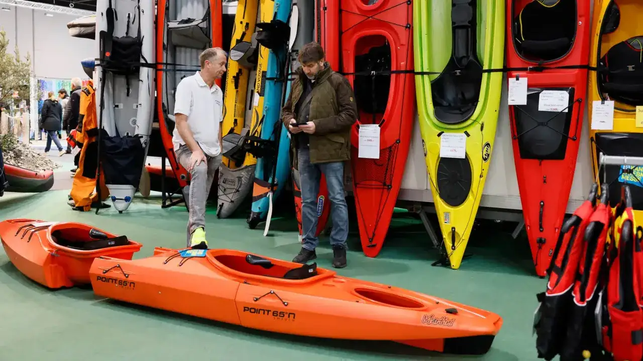 Zwei M&auml;nner betrachten Kajaks auf einer Messe. Ein orangefarbenes Kajak steht im Vordergrund. Viele bunte Kajaks sind im Hintergrund ausgestellt, bereit f&uuml;r das n&auml;chste Boot Programm.