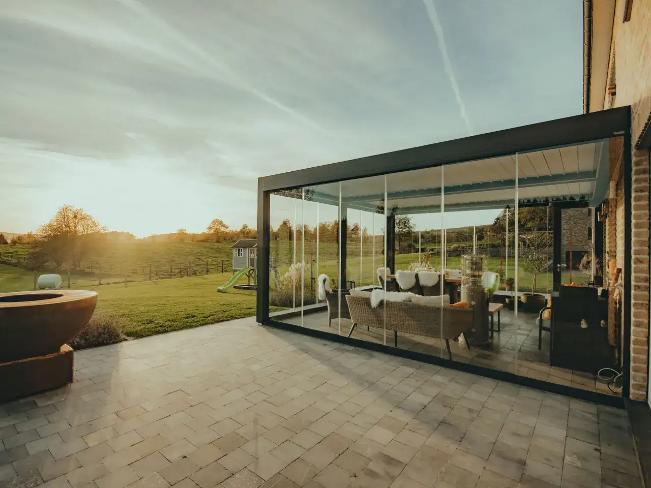 Un moderno sal&oacute;n acristalado, que es una p&eacute;rgola bioclim&aacute;tica, con muebles de exterior y vistas a un jard&iacute;n al atardecer.