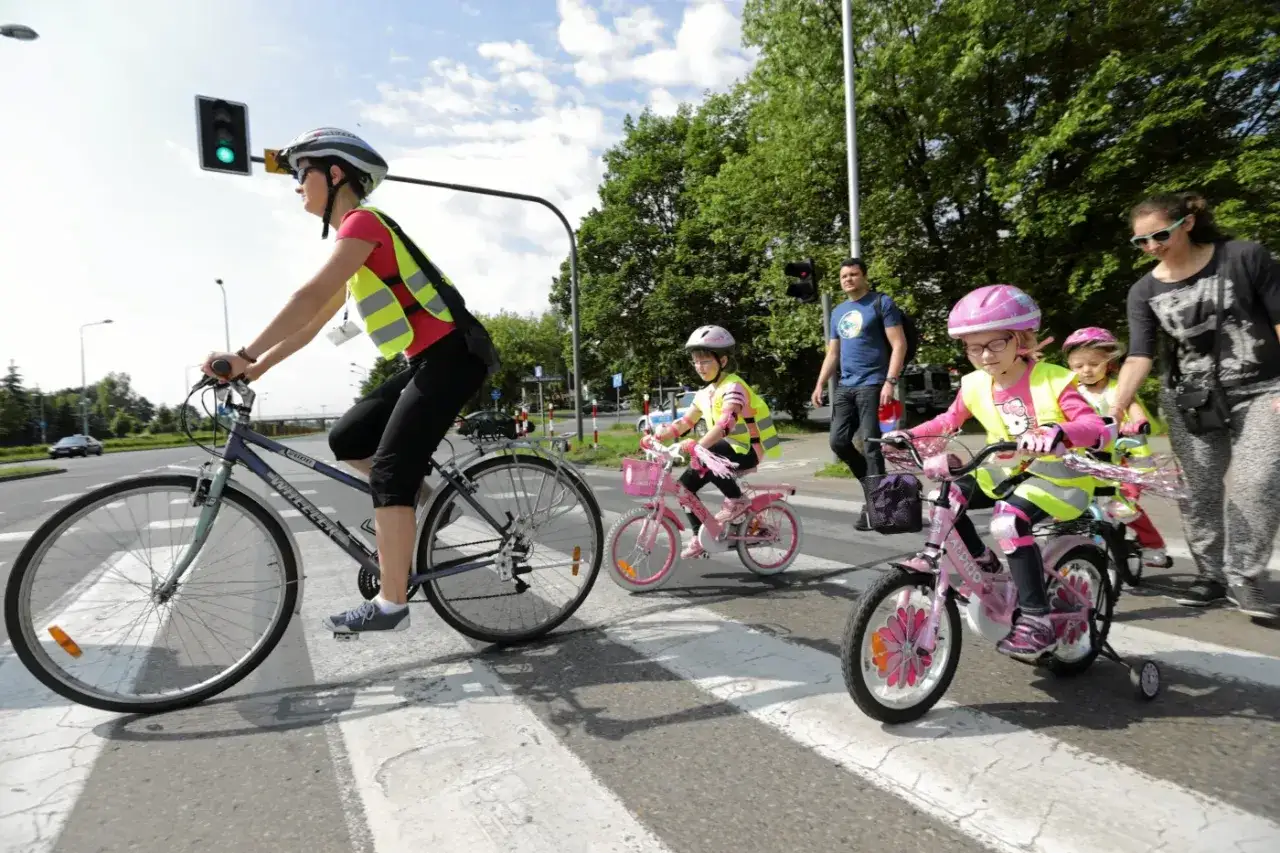 Rodzina na rowerach pokonuje przejście dla pieszych. Dzieci w kaskach i kamizelkach odblaskowych, mama i tata na drodze dla rowerów.