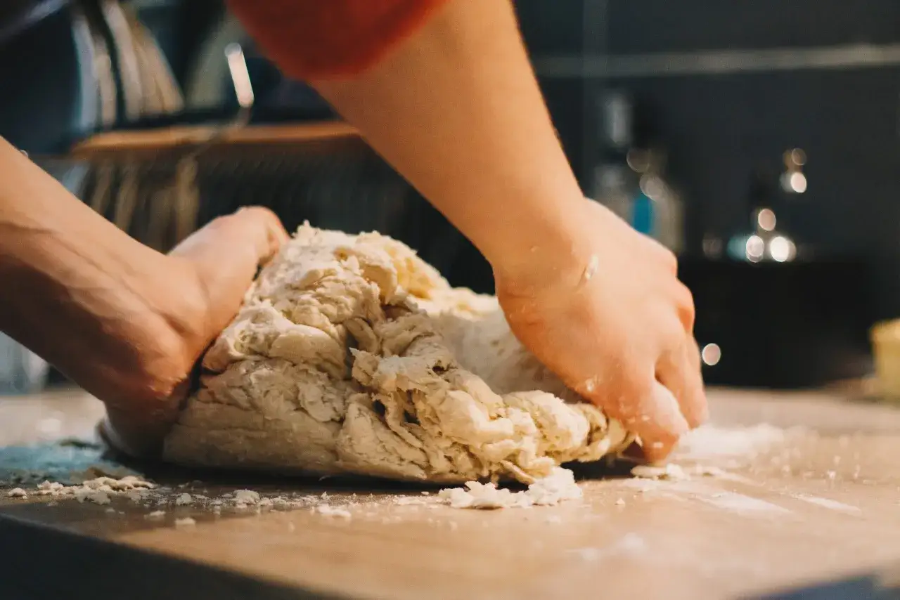 Mains p&eacute;trissant une p&acirc;te &agrave; pizza qui colle sur une planche en bois. La farine est saupoudr&eacute;e autour.