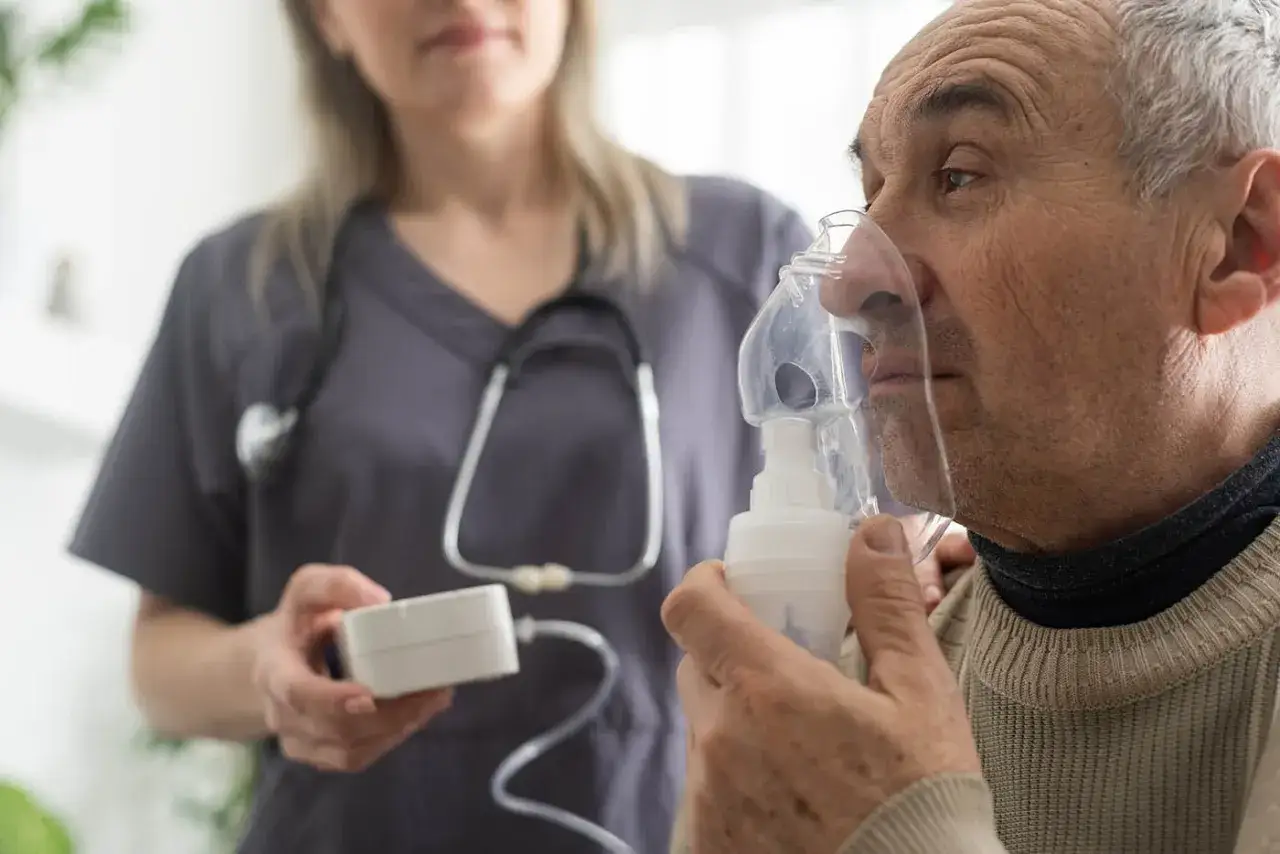 Mann mit Inhalator, ein Zeichen f&uuml;r COPD im Endstadium. Pflegekraft im Hintergrund.