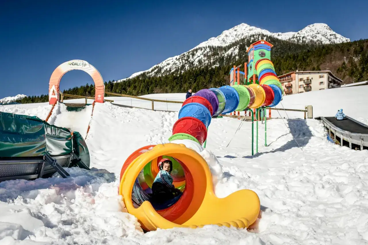 Bambino gioca nel colorato scivolo del **Pejo Kinderland** innevato, con montagne sullo sfondo.