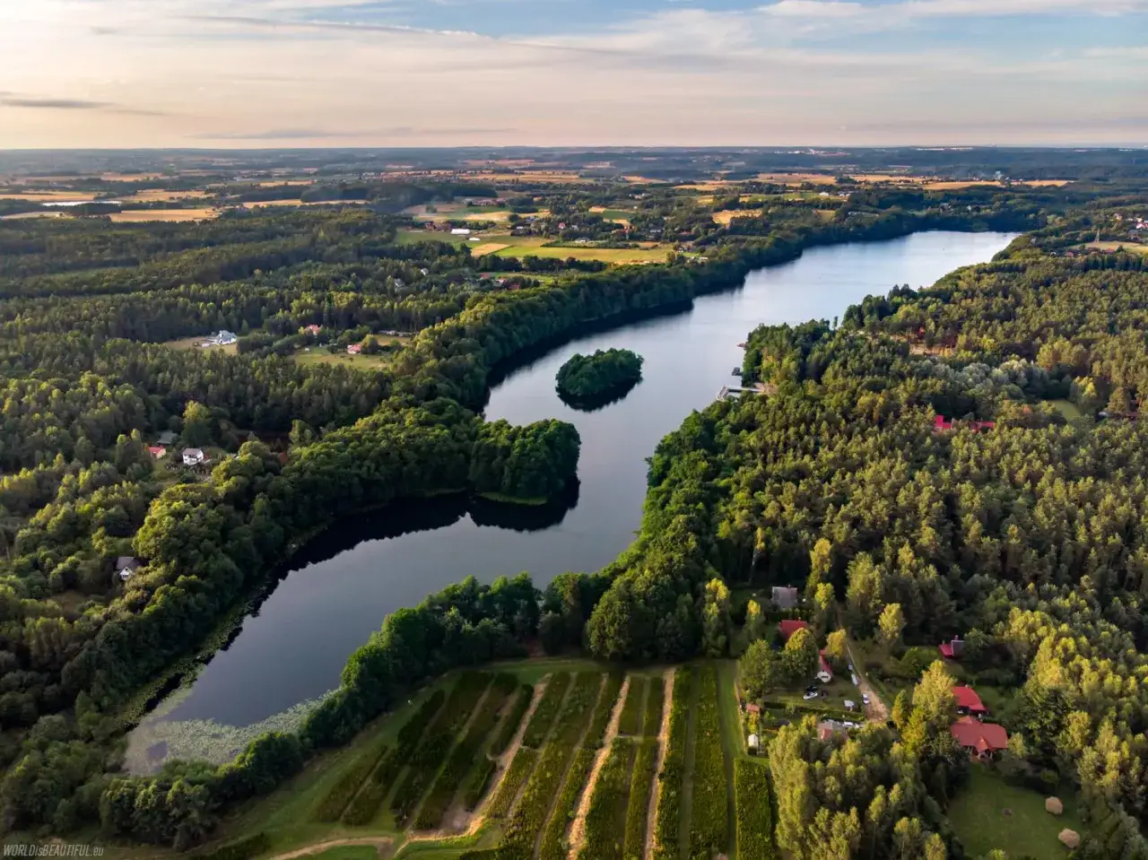 Malownicze jezioro otoczone lasem, z wyspą pośrodku. To piękne dzieło lodowca, gdzie widać pola uprawne i domy.