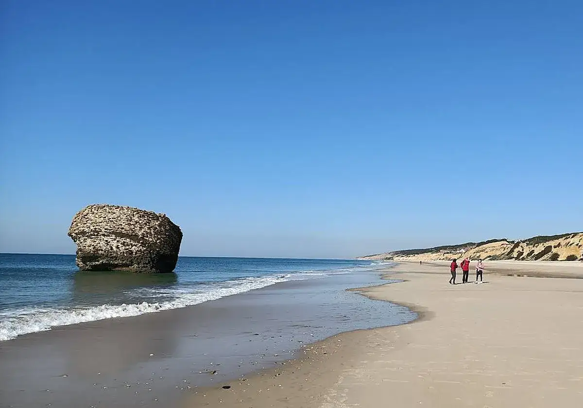Una roca solitaria emerge del mar en la playa de Castilla. Tres personas pasean por la arena dorada bajo un cielo azul despejado.