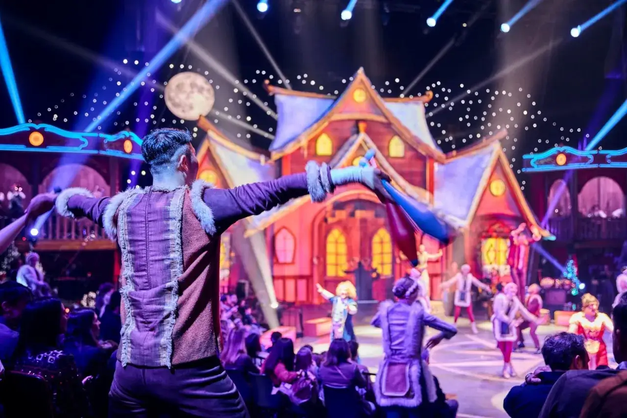 Un artista hace malabares con mazas de colores en un escenario iluminado, parte de un vibrante circo para ni&ntilde;os con una casa de cuento de hadas de fondo.