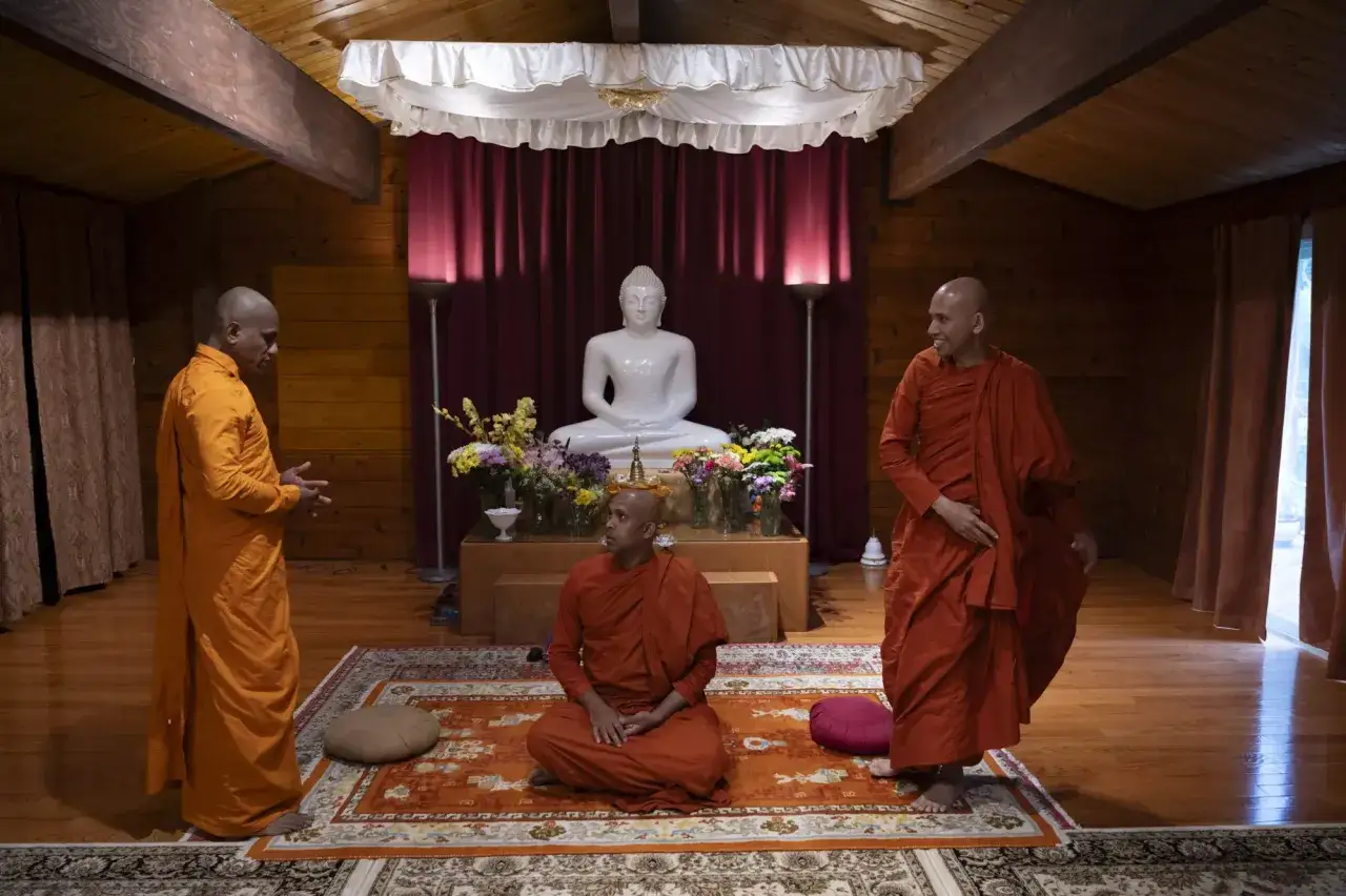 Three monks in orange robes gather in a serene room with a Buddha statue. Their conversation echoes timeless monk sayings.