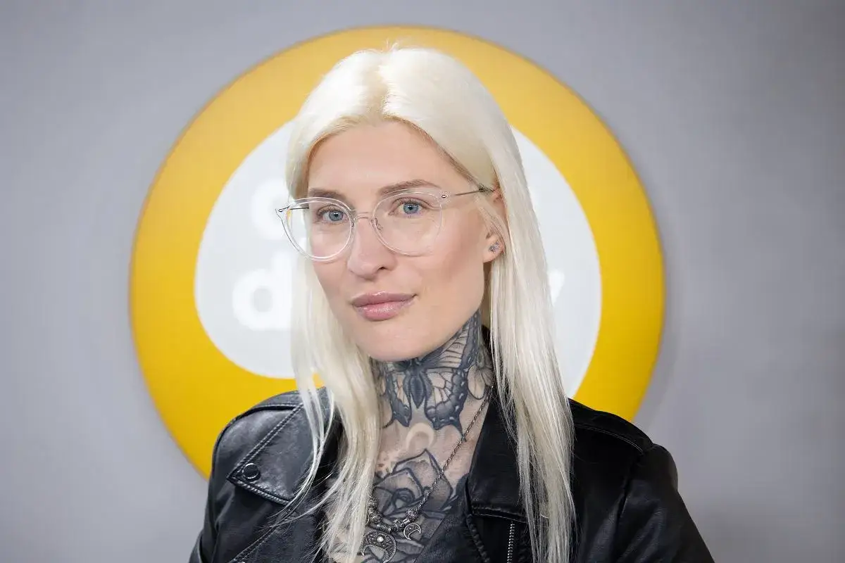 Natalia Sisik w okularach i skórzanej kurtce, z tatuażami na szyi, pozuje na tle żółto-białego koła.