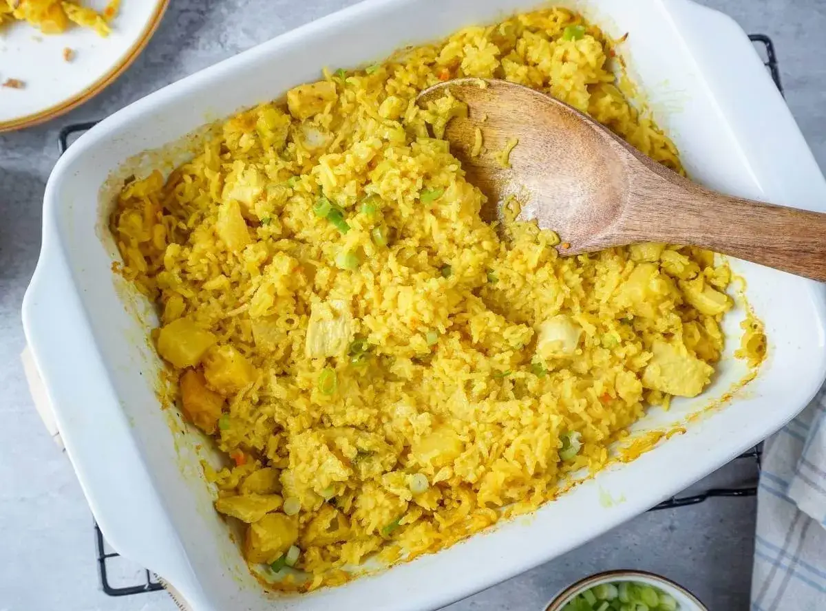 Ein gelber Reisauflauf mit Ananasstücken und Hühnchen wird in einer weißen Auflaufform serviert. Ein Holzlöffel liegt darin. Perfekt, um Reis im Ofen zu kochen.