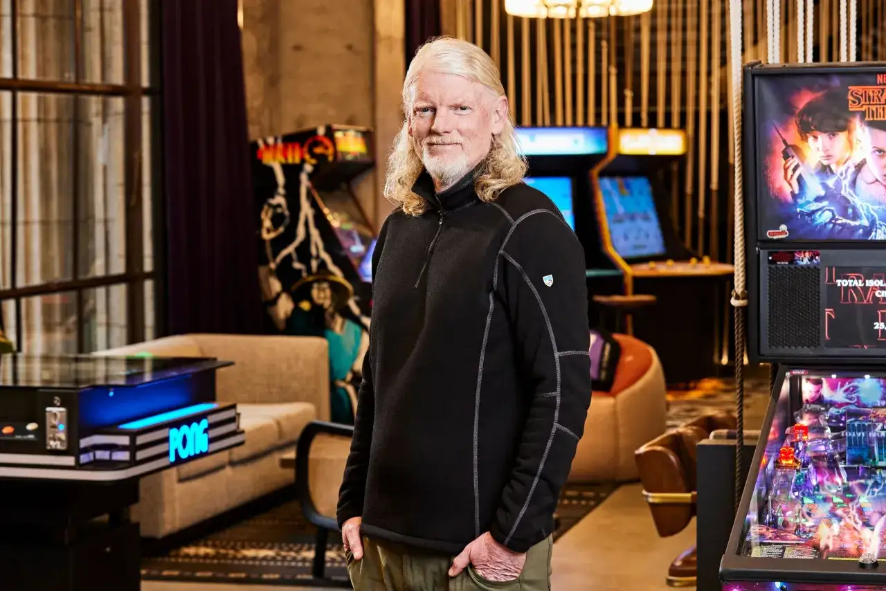 El CEO de Twitch, con su característico cabello rubio, posa sonriente rodeado de máquinas arcade y un pinball de Stranger Things.