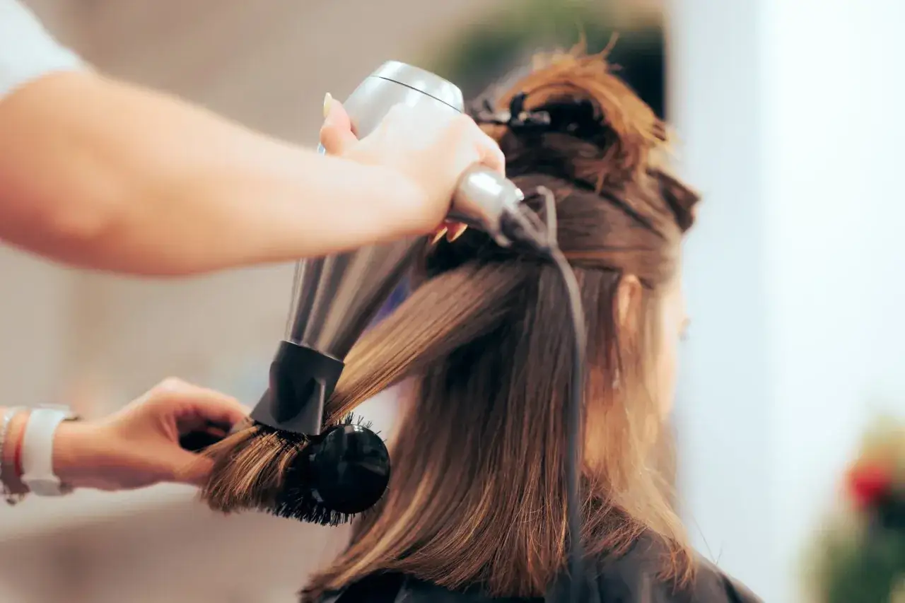 Profesjonalny styling włosów w salonie. Fryzjerka suszy i modeluje włosy klientki okrągłą szczotką i suszarką.
