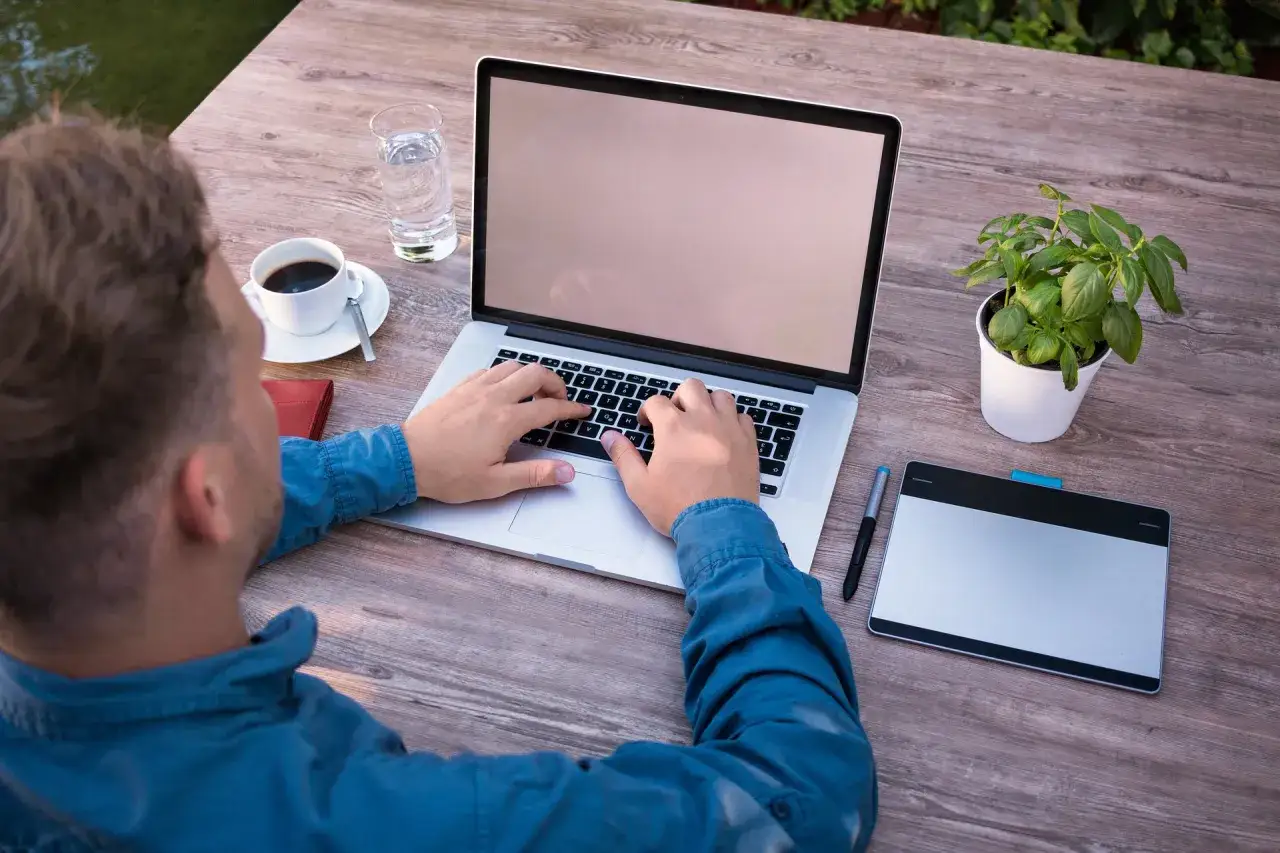 Mann tippt auf Laptop, Kaffee, Wasser, Pflanze. Der Weg, um **freelancer werden** zu können, beginnt oft so.