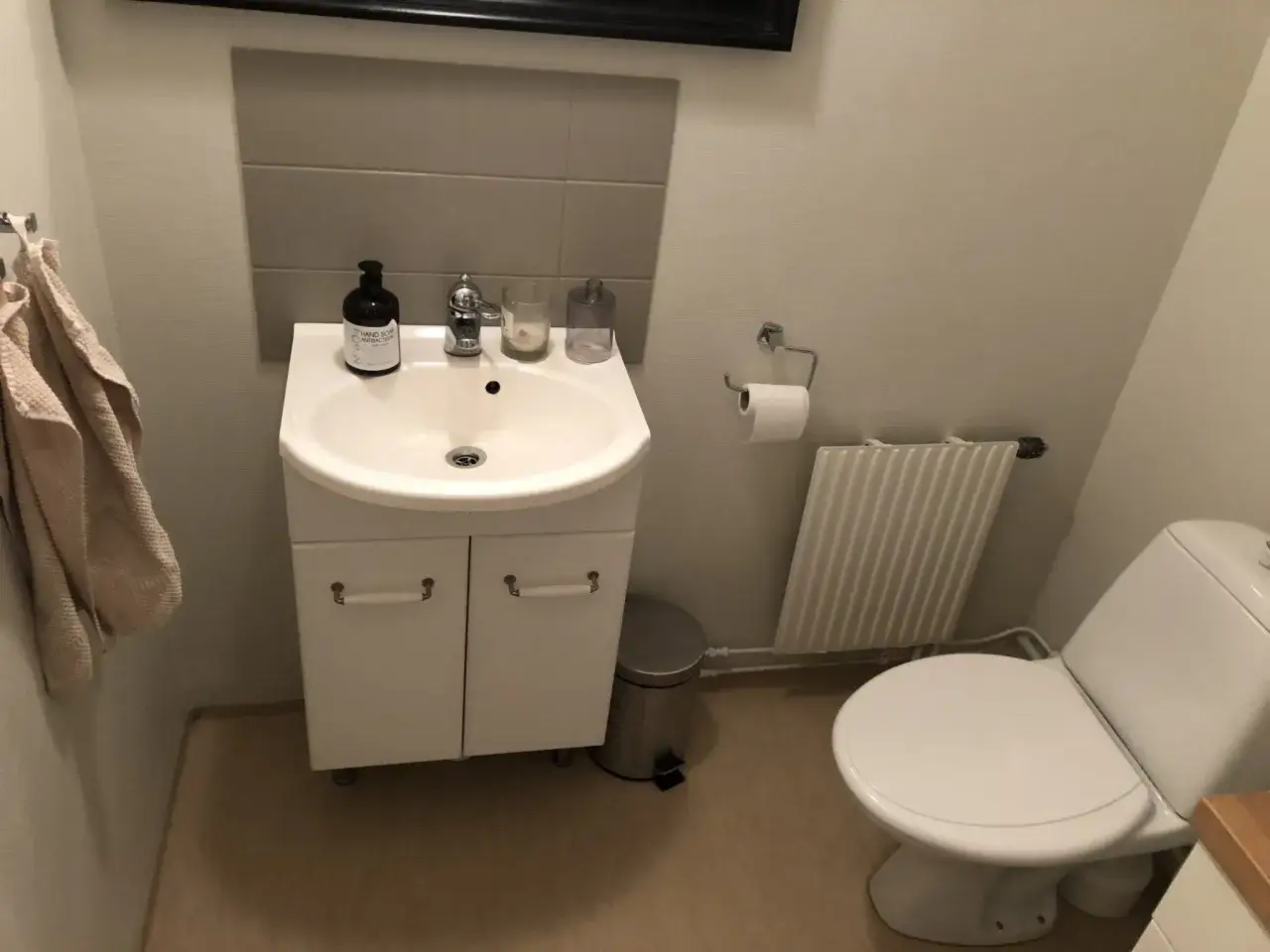 Ett litet badrum med handfat, toalett och en väggmonterad handdukshängare. Golvet ser ut att vara en våtrumsmatta som kan behöva målas om.