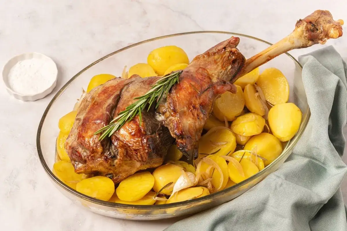 Pierna de cordero al horno dorada, servida con patatas asadas, romero y ajo en una fuente de cristal.