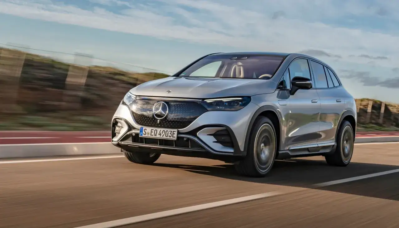 Un elegante modelo Mercedes plateado, un SUV el&eacute;ctrico, avanza por una carretera con un cielo azul y nubes.