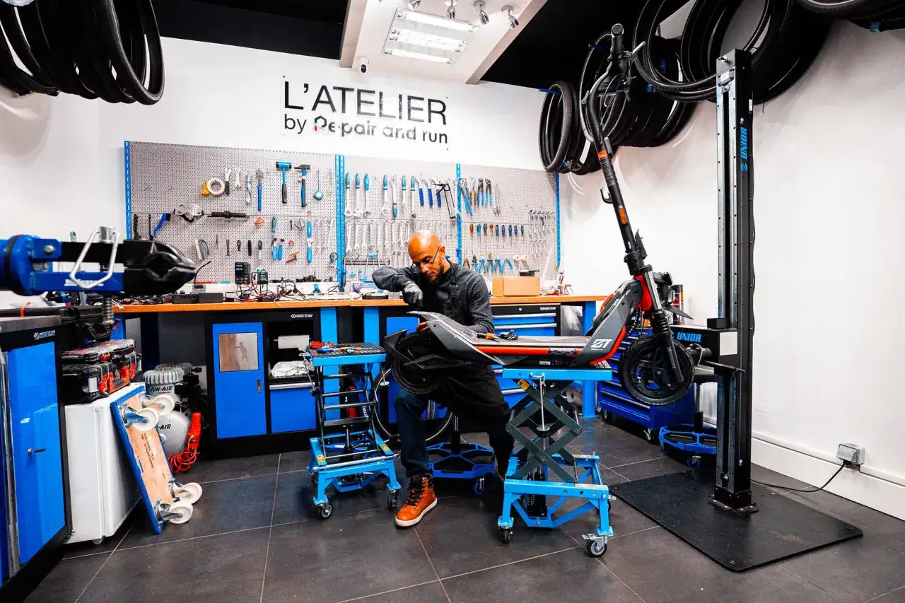 Un mécanicien répare une trottinette électrique dans un atelier bien équipé. Des outils sont accrochés au mur, des pneus sont suspendus au plafond.