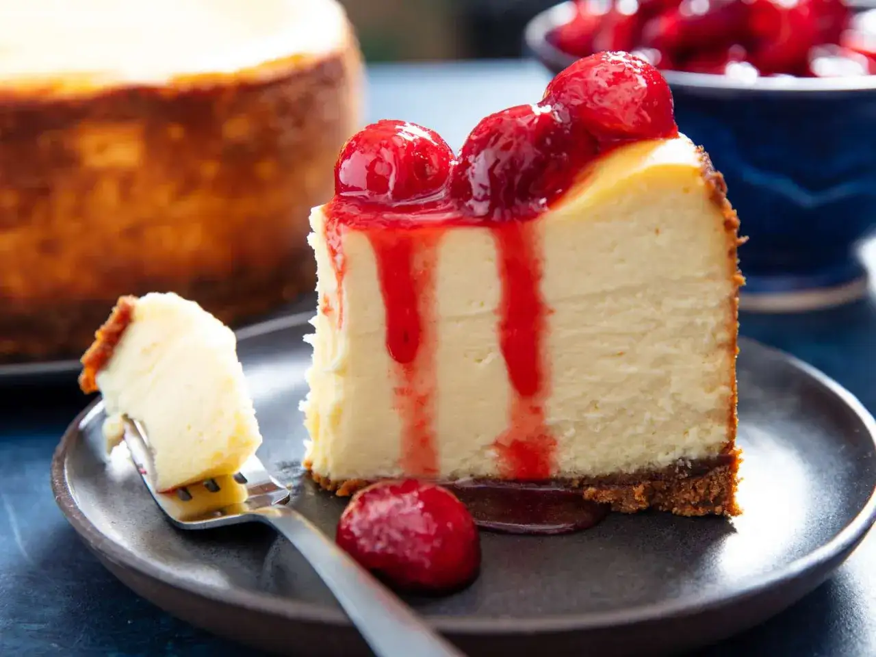 Ein Stück cremiger american cheesecake mit keksboden, garniert mit roten Beeren und Sauce. Ein weiterer Kuchen ist im Hintergrund zu sehen.