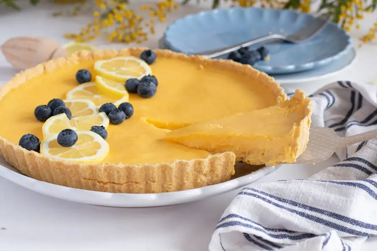 Tarta de limón cremosa, decorada con rodajas de limón y arándanos frescos. Una porción se levanta con una espátula, lista para ser disfrutada.