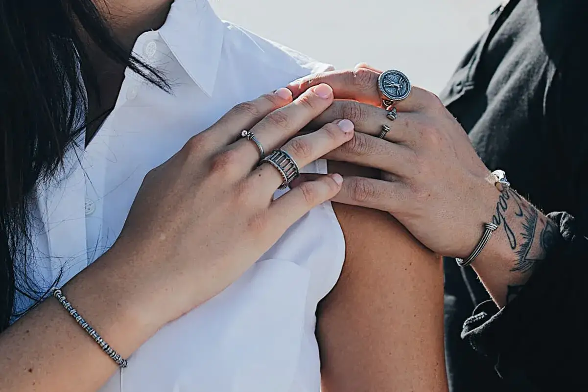 Mani con anelli, uno con una bussola, un altro a gabbia. Il bracciale al polso tatuato suggerisce dove si mette l'anello da single.
