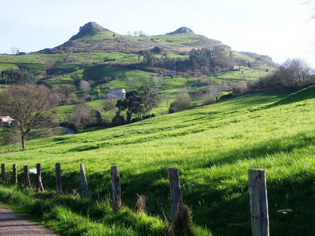 Verde prado en la ruta Tetas de Liérganes, con un camino rural y un cercado de madera. Montañas al fondo.