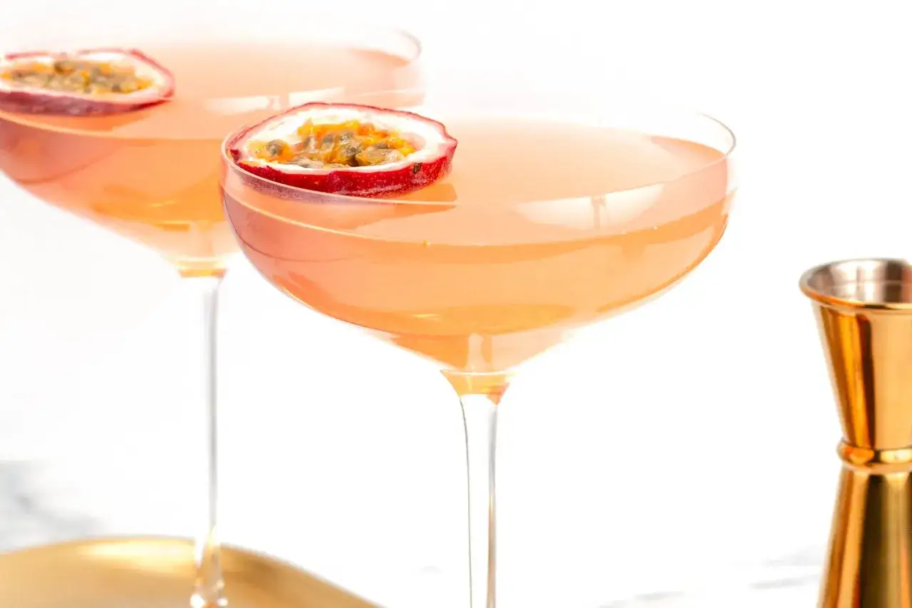 Dwa kieliszki z drinkiem Pornstar Martini, ozdobione połówkami marakui. Obok złoty jigger.
