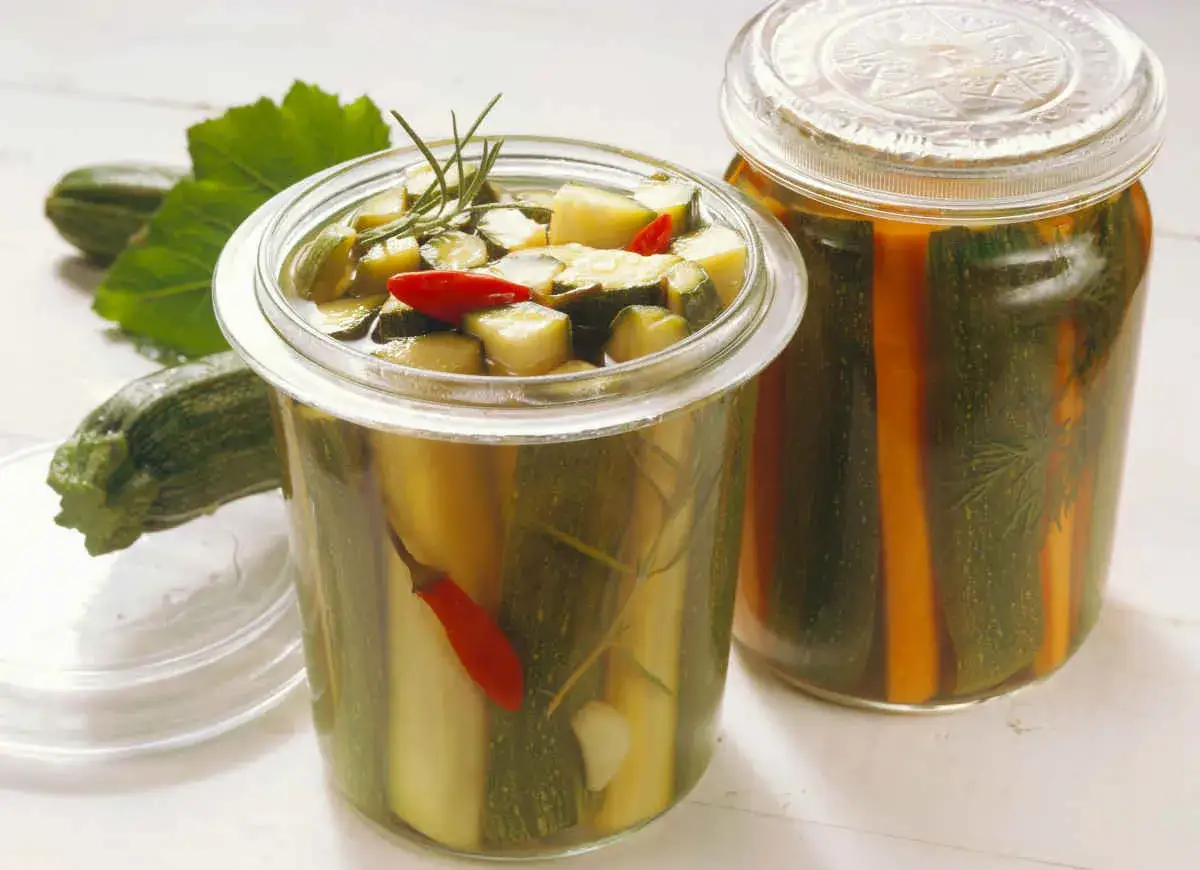 Słoiki z cukinią marynowaną po włosku, z dodatkiem chili i ziół. Obok leżą świeże cukinie i liście.