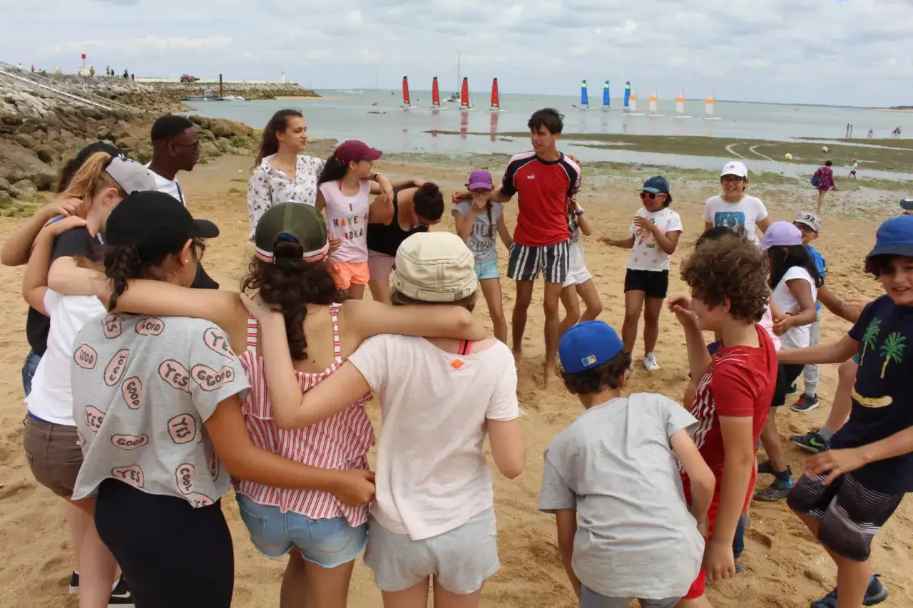 Une joyeuse colonie à la mer, des enfants rient et s'amusent sur la plage, des voiliers naviguent au loin.