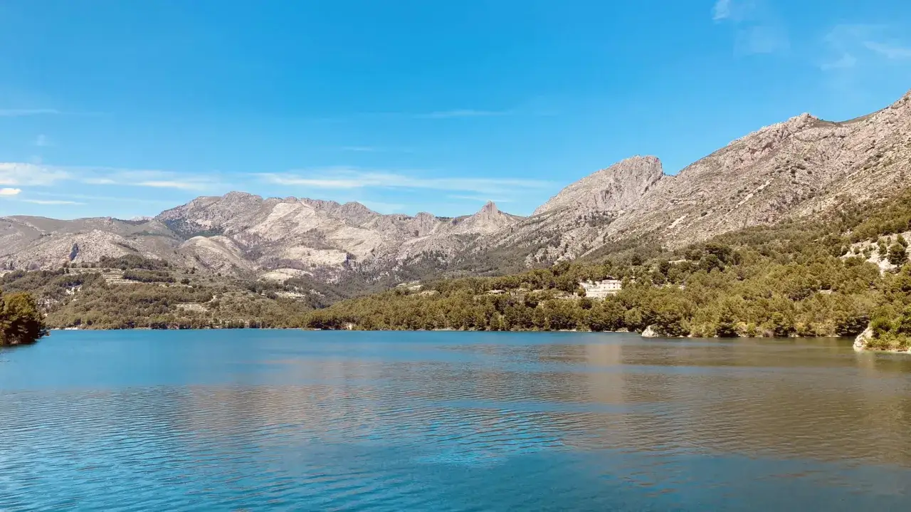 Blauer See vor bergiger Landschaft mit dem Castell de Guadalest in der Ferne.