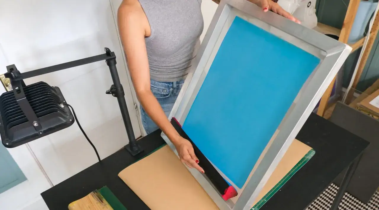 Persona prepara un marco de serigrafía con una rasqueta para el proceso de impresión.