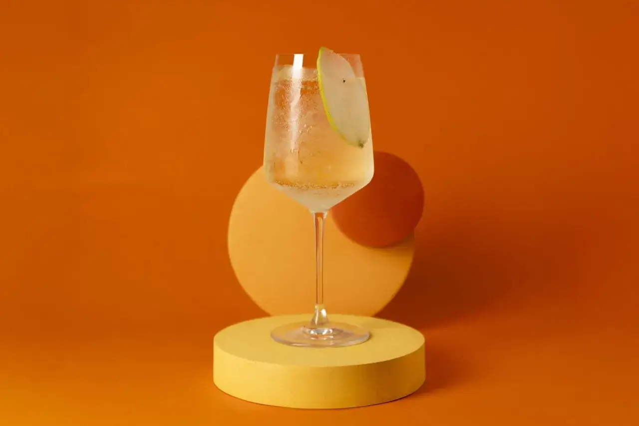 Ett glas med en uppfriskande drink med Xante, garnerad med en tunn p&auml;ronklyfta. St&aring;r p&aring; en gul piedestal mot en orange bakgrund.