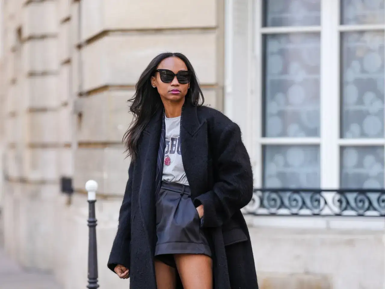 Frau in schwarzem Mantel und lässigem T-Shirt, die zeigt, wie man ledershorts kombinieren kann. Sonnenbrille und dezentes Make-up runden den Look ab.