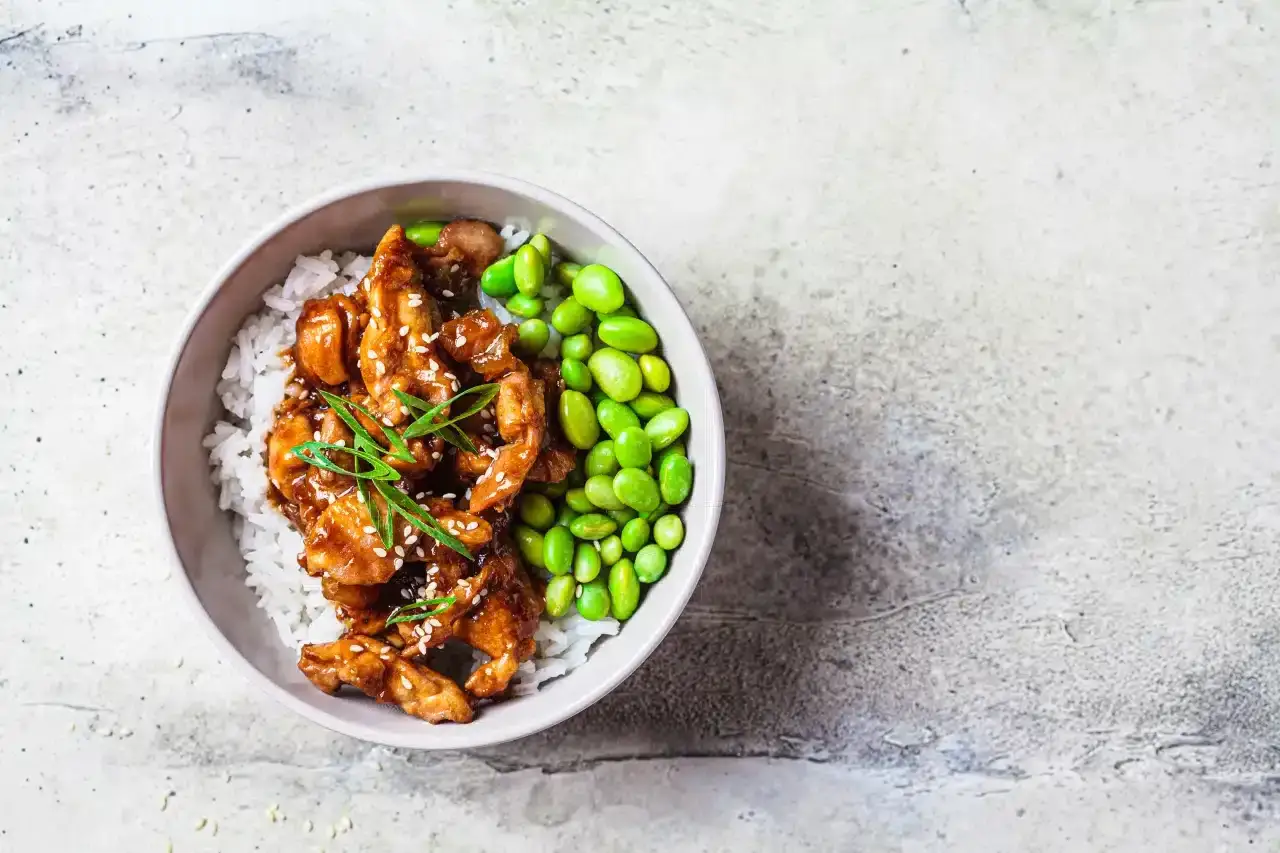 Apetyczny bowl z kurczakiem w sosie teriyaki, ryżem i zielonymi fasolkami edamame, posypany sezamem i szczypiorkiem.