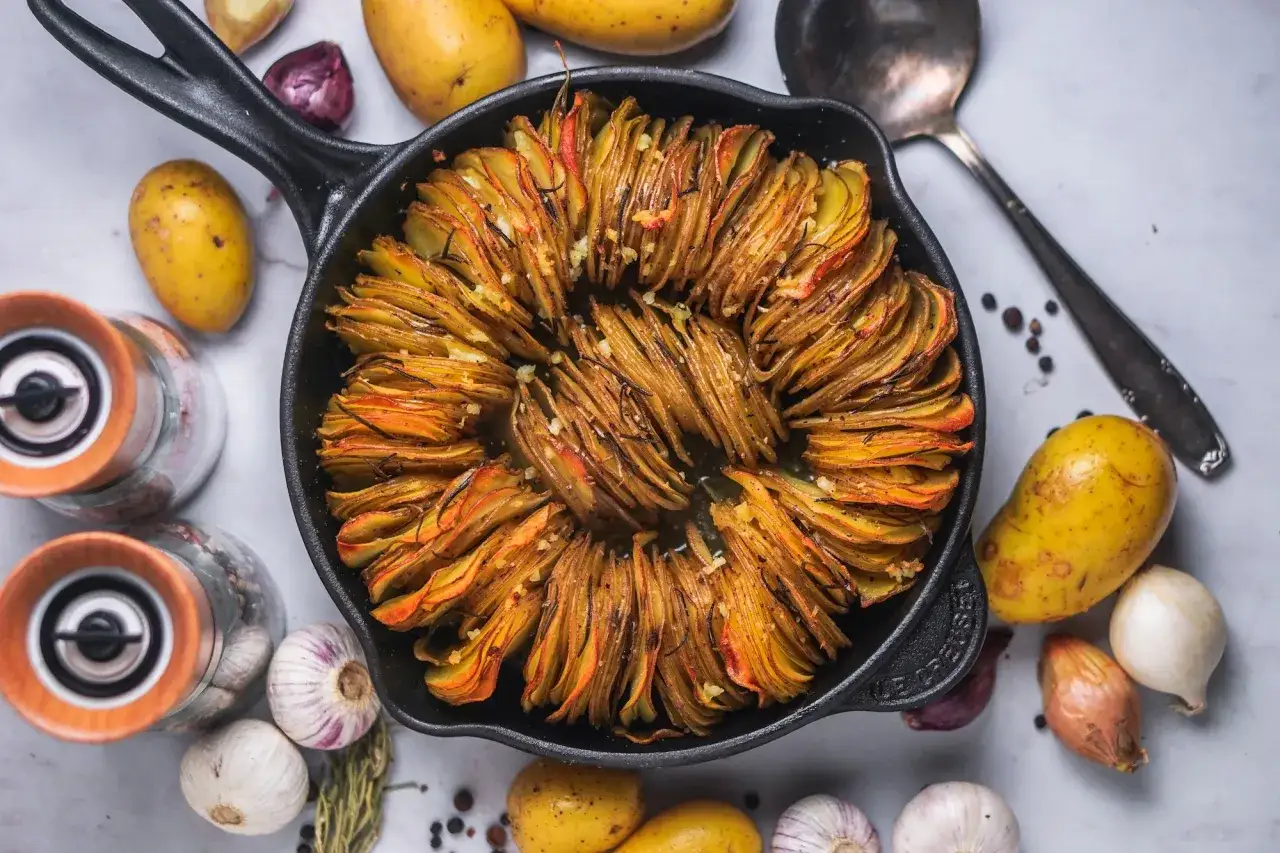 Knoblauch Kartoffeln in einer Gusseisenpfanne, umgeben von frischen Kartoffeln, Knoblauch und Zwiebeln.